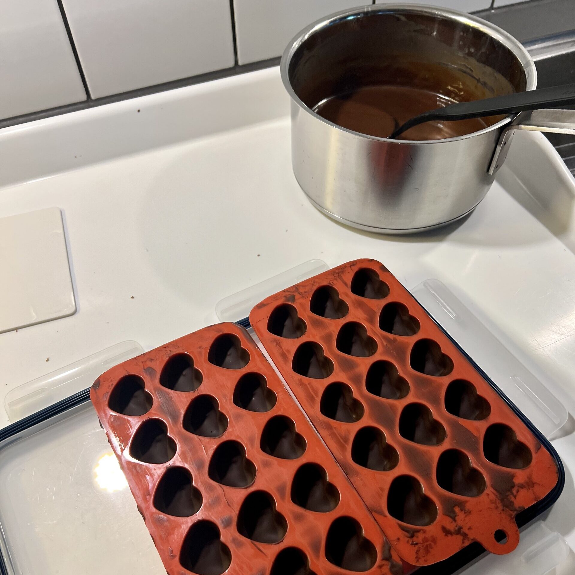 Röda hjärtformade pralinformar fyllda med mörk stelnad choklad i förgrunden, kastrull med kaffefyllning och en svart slev i bakgrunden.